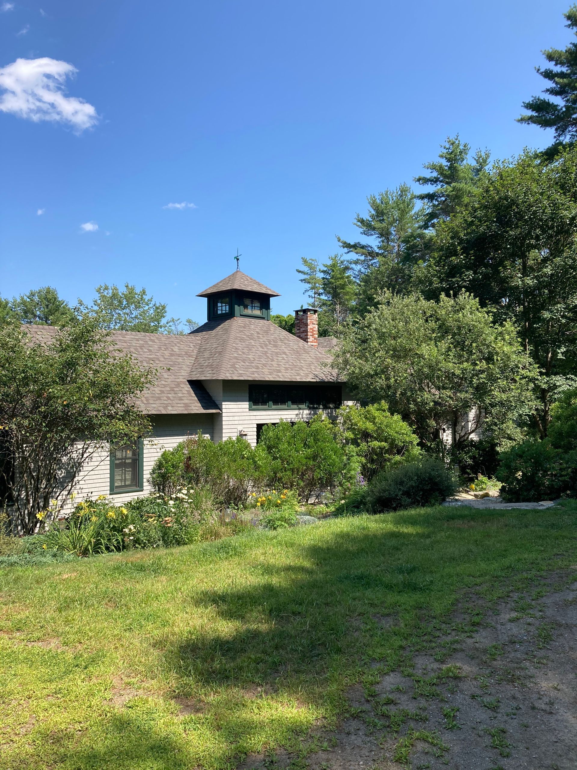 A large white house with a gray roof is surrounded by trees and grass on a sunny day.