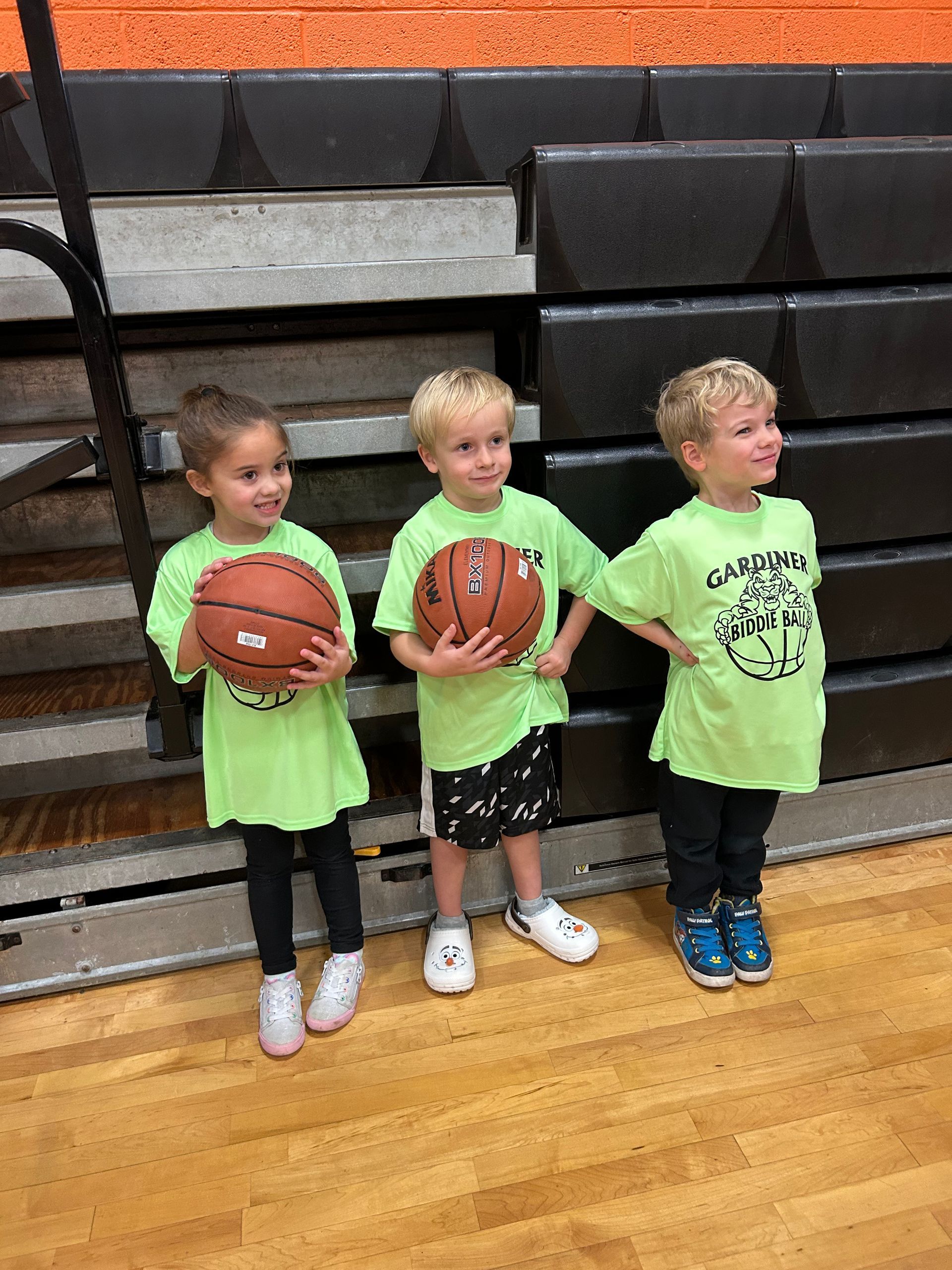 Three young children are standing on a basketball court holding basketballs.