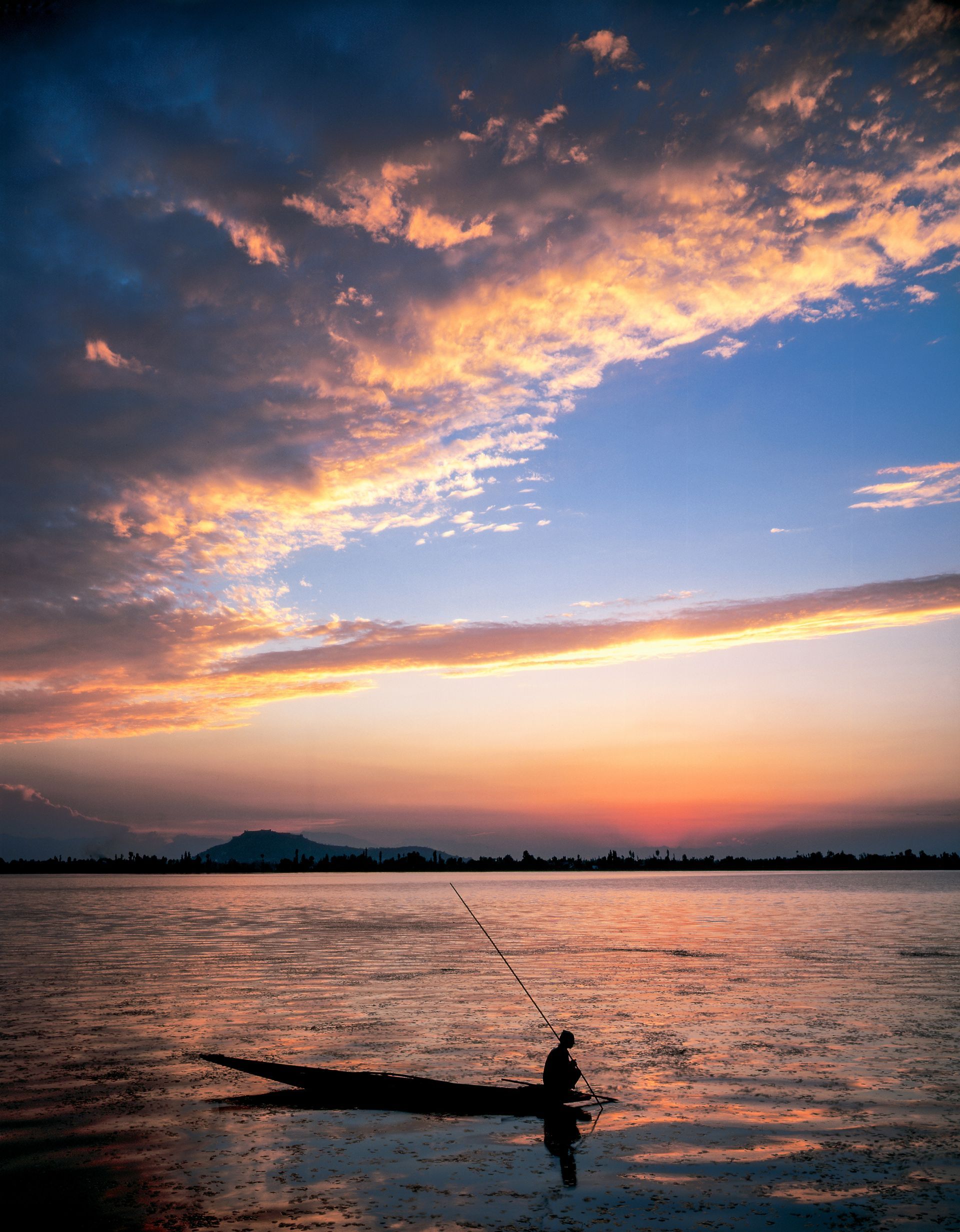 A man is fishing in a boat on a lake at sunset.