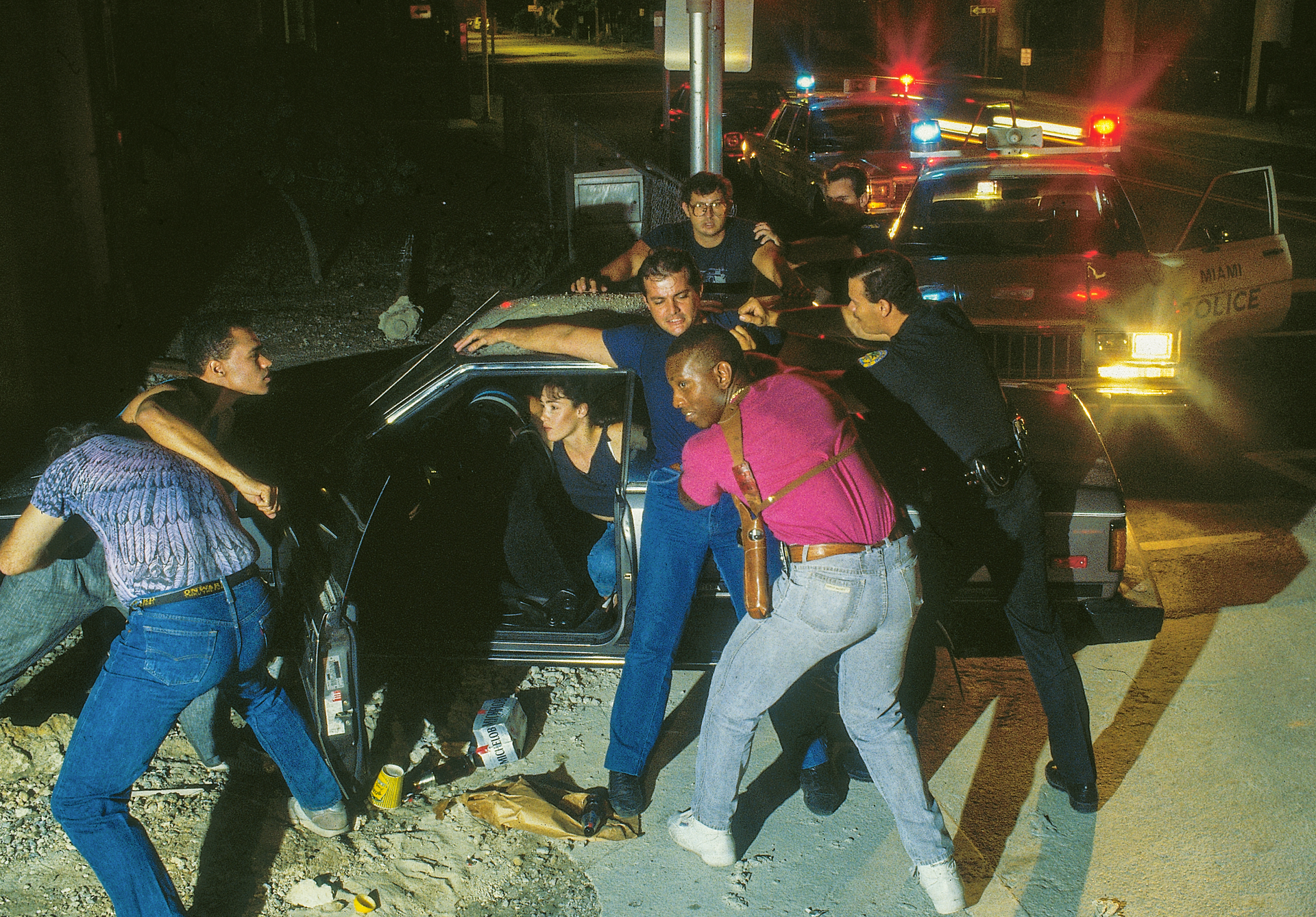 A group of people standing around a car with a police car in the background