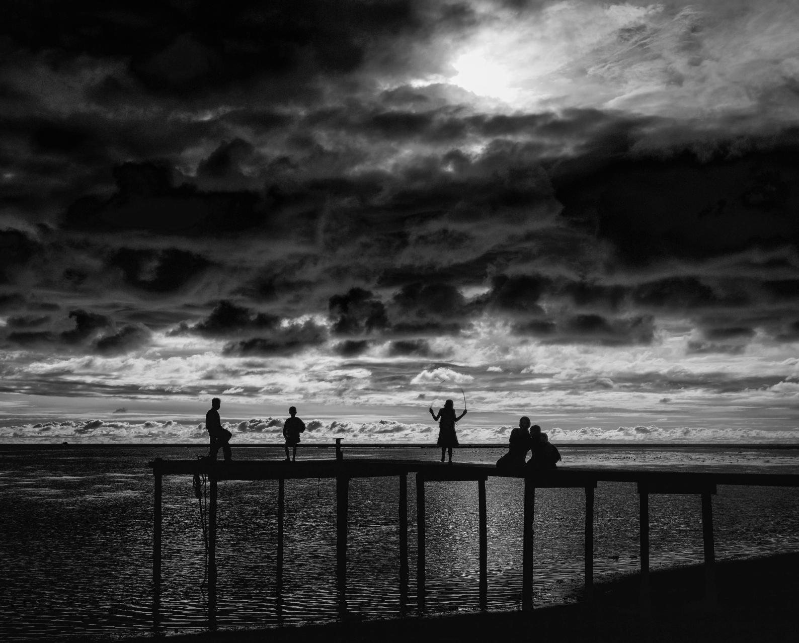 A black and white photo of people standing on a pier overlooking the ocean.