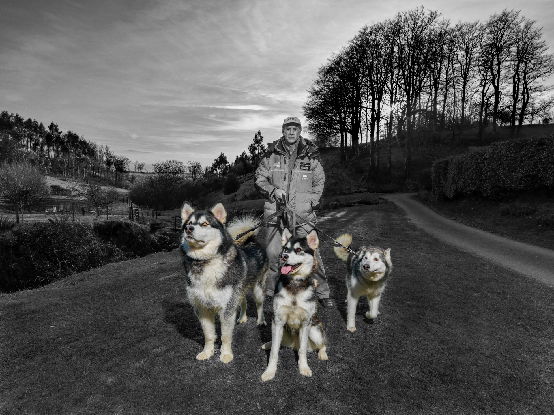 A man is standing next to three husky dogs on a dirt road.