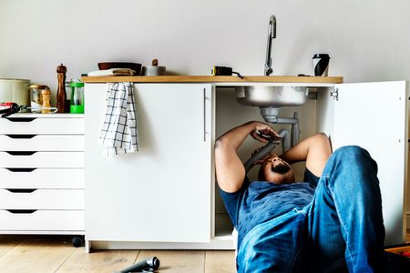 MDM Plumbing & Sewer LLC | A man is laying on the floor fixing a sink in a kitchen.