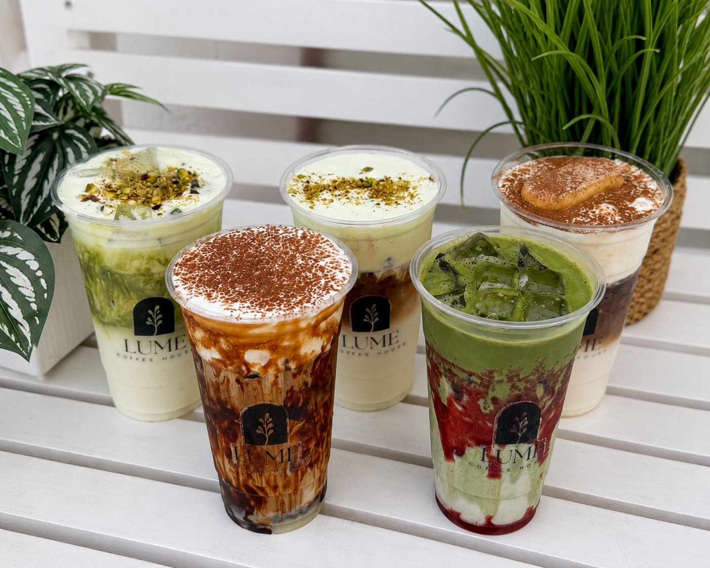 Five colorful iced drinks in clear cups on a white bench, topped with froth, herbs, and cocoa powder