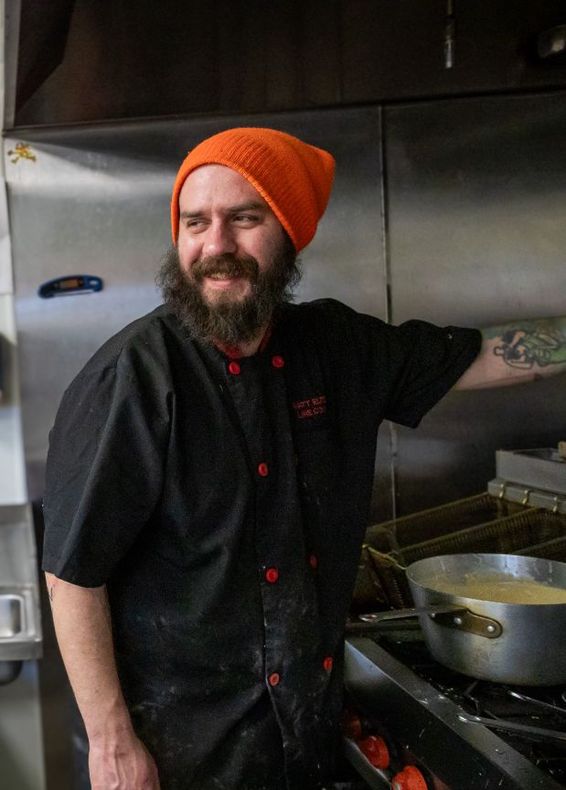 A man with a beard and an orange hat is standing in front of a stove in a kitchen.