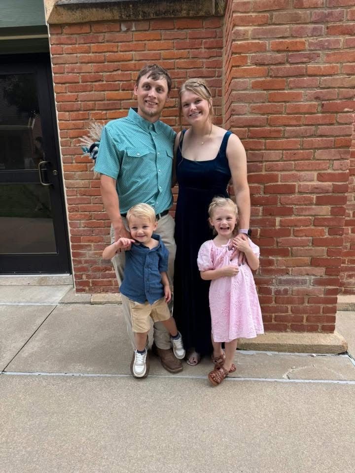 Family of four poses in front of a brick building. Man in green shirt, woman in black dress, and two young children.