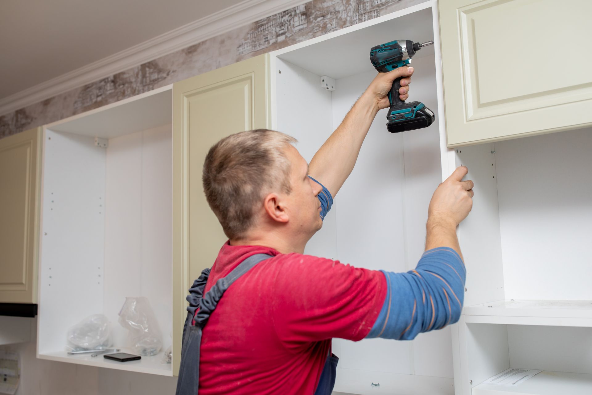 Man in red shirt installing a cabinet with a drill in a kitchen.