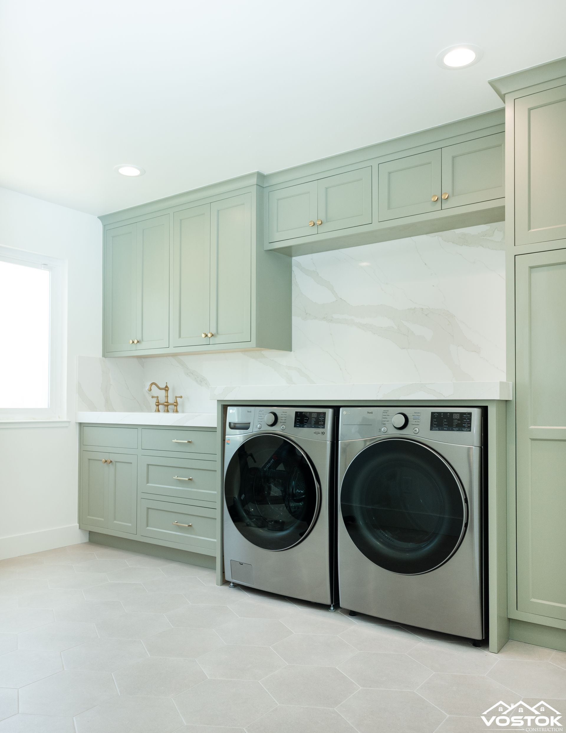 Modern laundry room featuring custom cabinetry, sleek white countertops, marble-patterned backsplash, and two front-loading washing machines set on light hexagonal tile flooring.