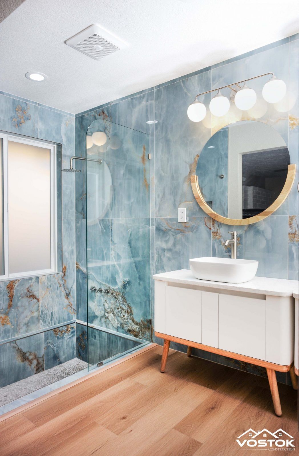 A bathroom with blue tiles , a sink , a mirror and a walk in shower.