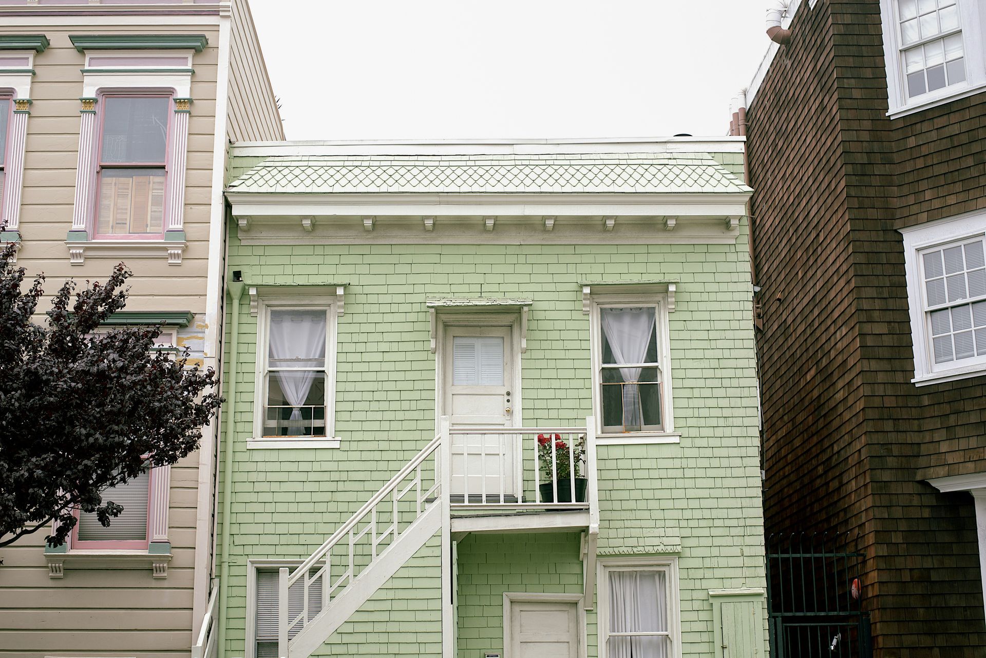 Green two-story house, white trim, balcony flowers, staircase, neighboring buildings.