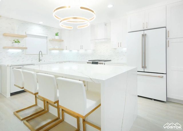Modern white kitchen with marble island, gold-framed bar stools, and sculptural pendant lighting