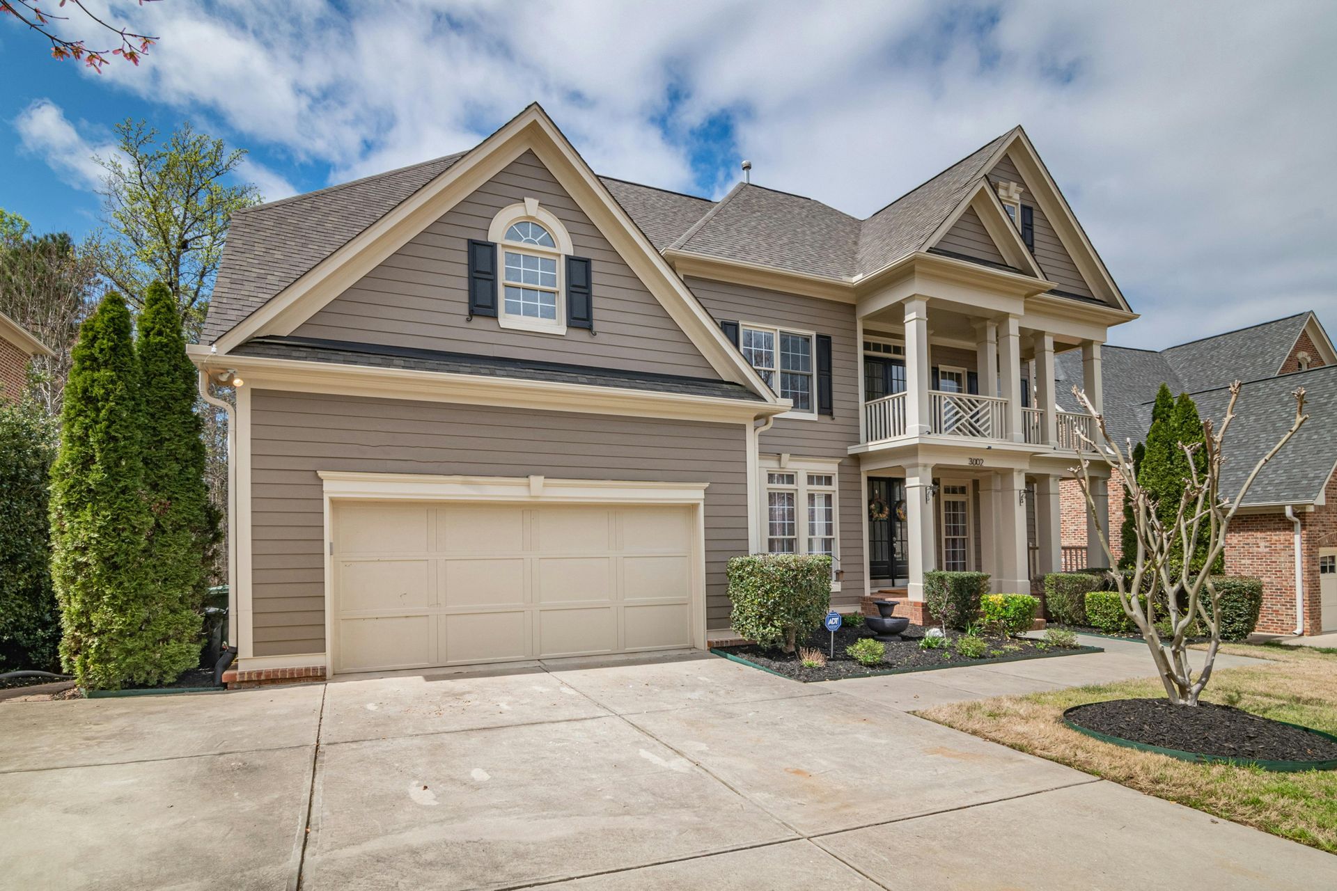 Two-story beige house with gabled roof, porch, and landscaped front yard.