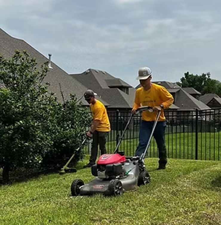Two people mowing a lawn, one with a mower, the other trimming near a bush, wearing yellow shirts.