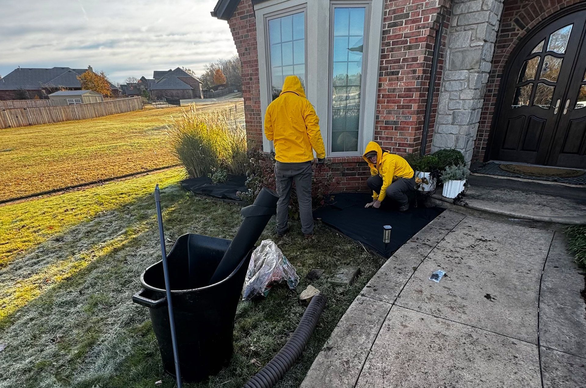 Two people in yellow jackets install landscaping near a brick house with a window and a stone archway; tools are nearby.