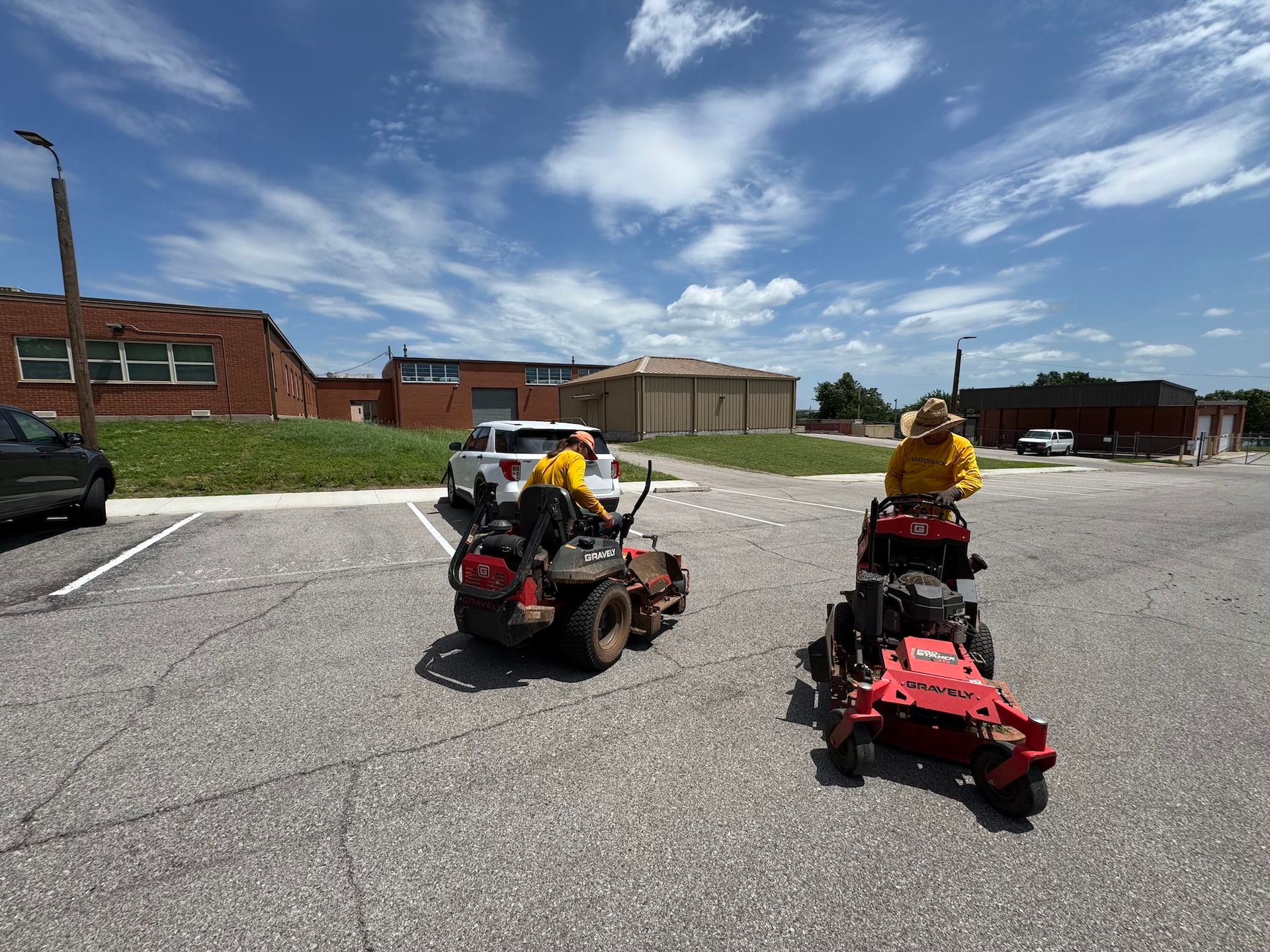 Two people on red lawnmowers in a parking lot. They are wearing yellow shirts, mowing on a sunny day.