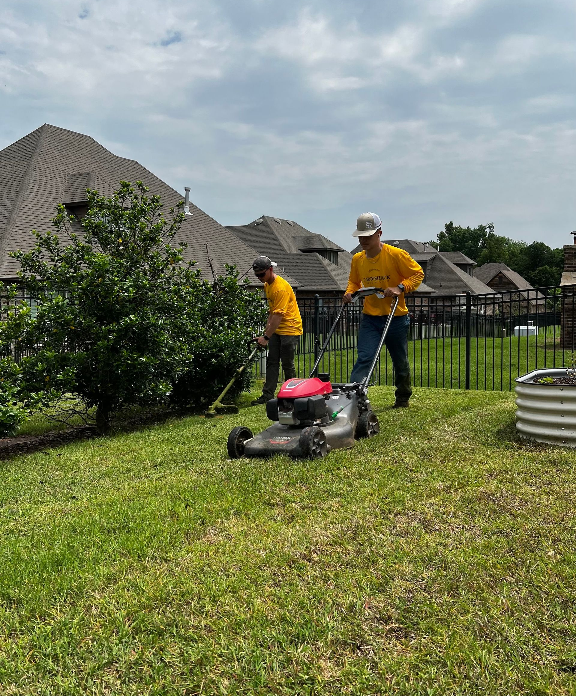 Two people in yellow shirts mowing a grassy yard in front of houses.