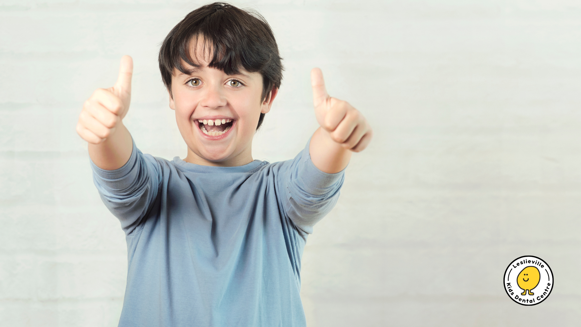Smiling child with dark hair gives thumbs up, wearing a blue shirt, against a white background.