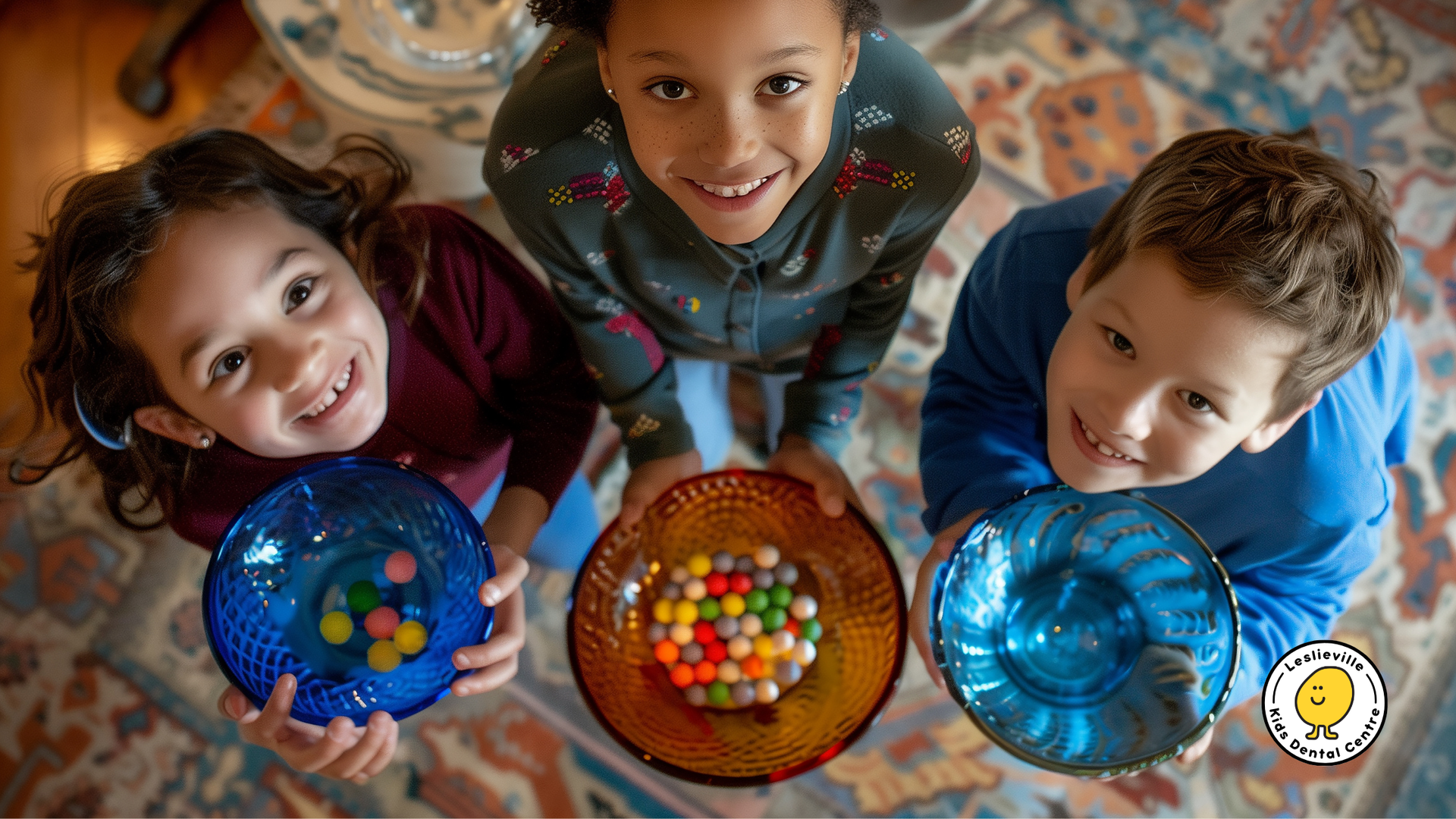Three smiling children holding colored bowls of candy, looking up.