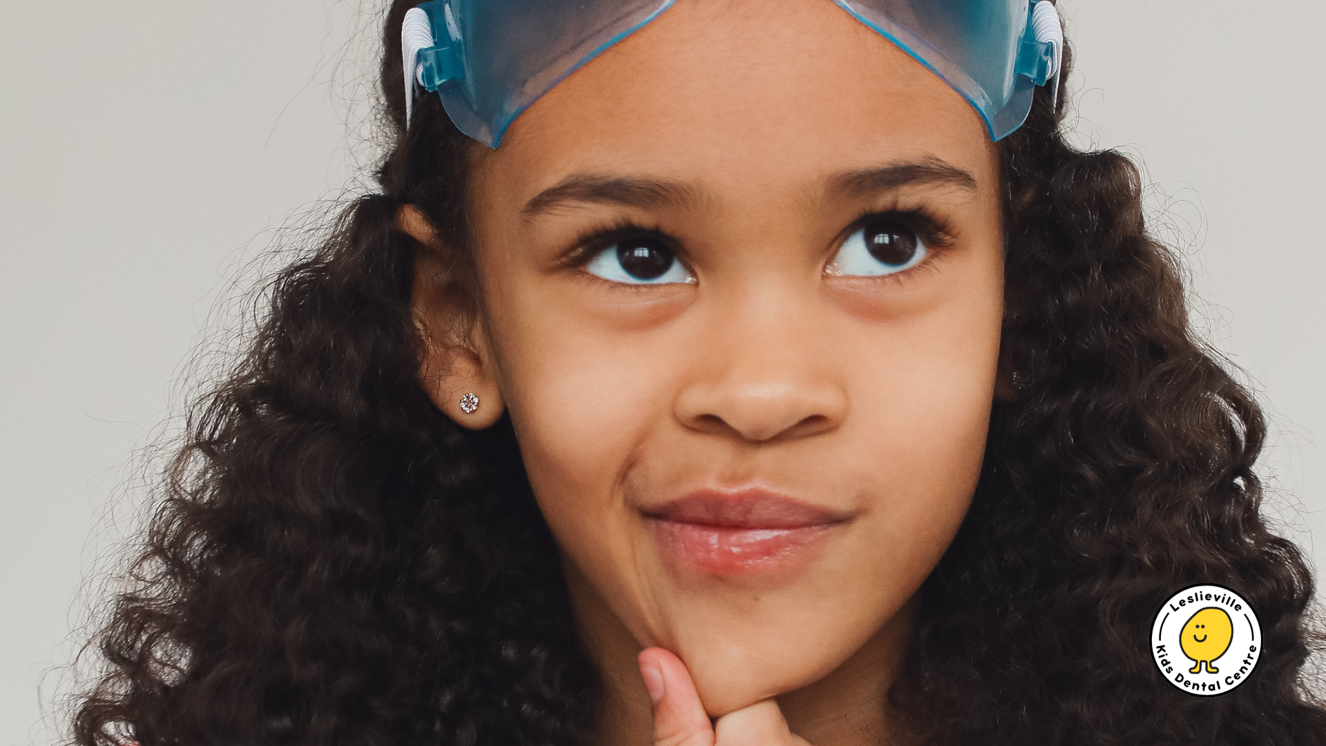 Girl with curly hair wearing goggles, looking up thoughtfully, hand on chin.