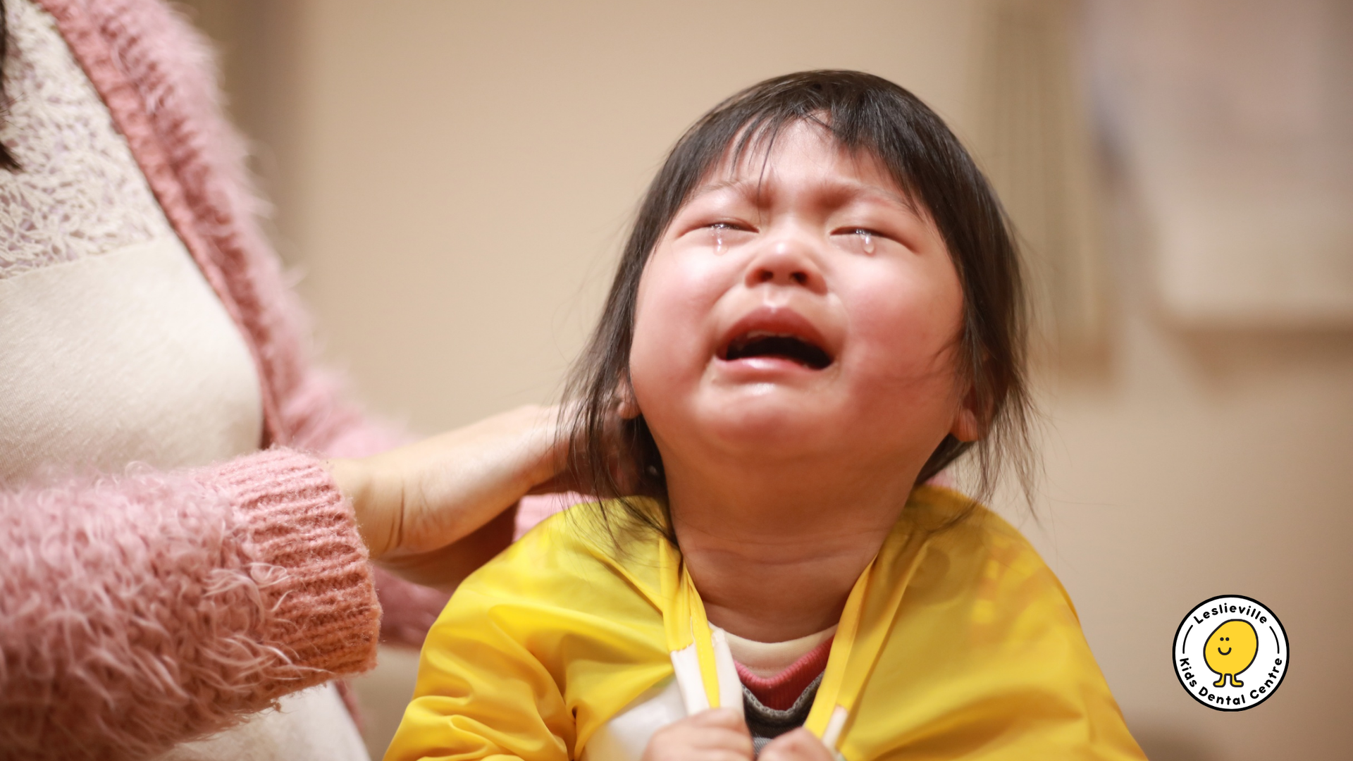 A crying young child in a yellow coat has tears on their face, being comforted by a hand on their shoulder.