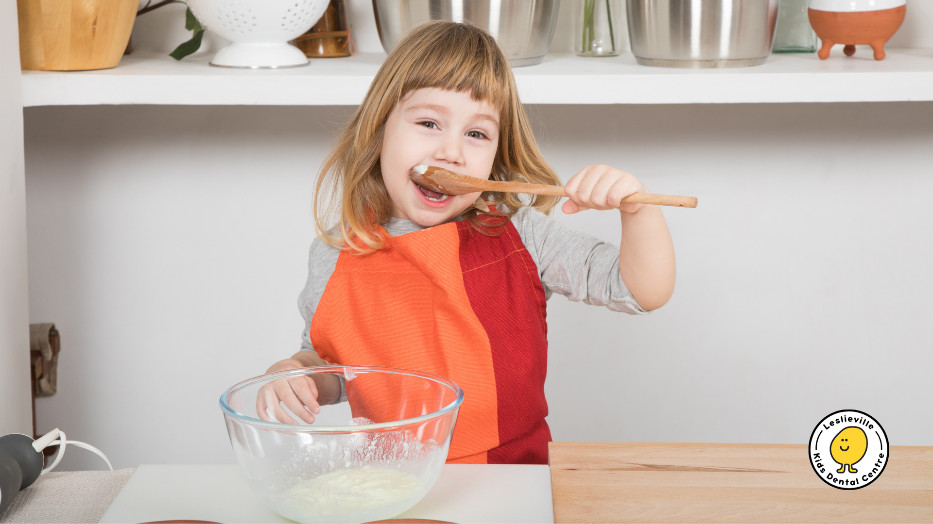 Child tasting batter from a wooden spoon in a kitchen, wearing an apron.