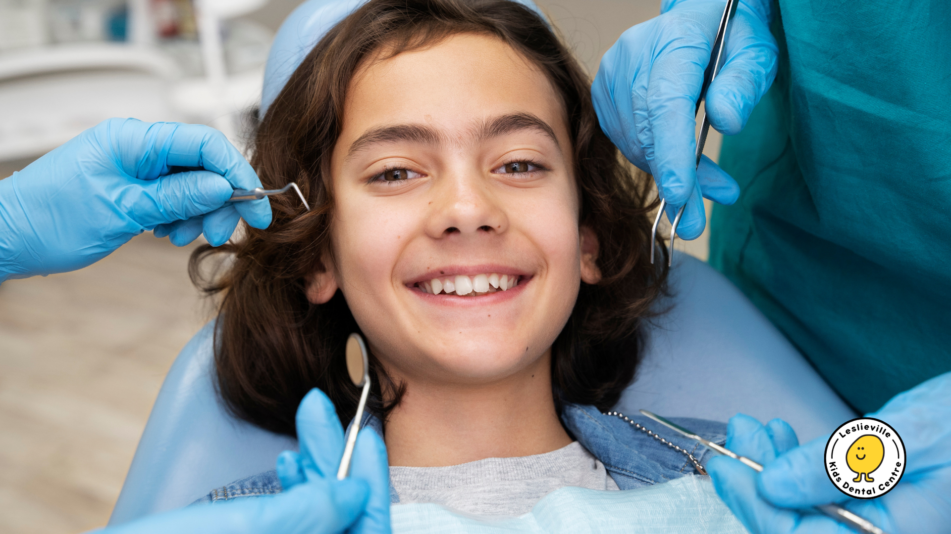Smiling child in a dentist chair with dental tools held by gloved hands surrounding their face.