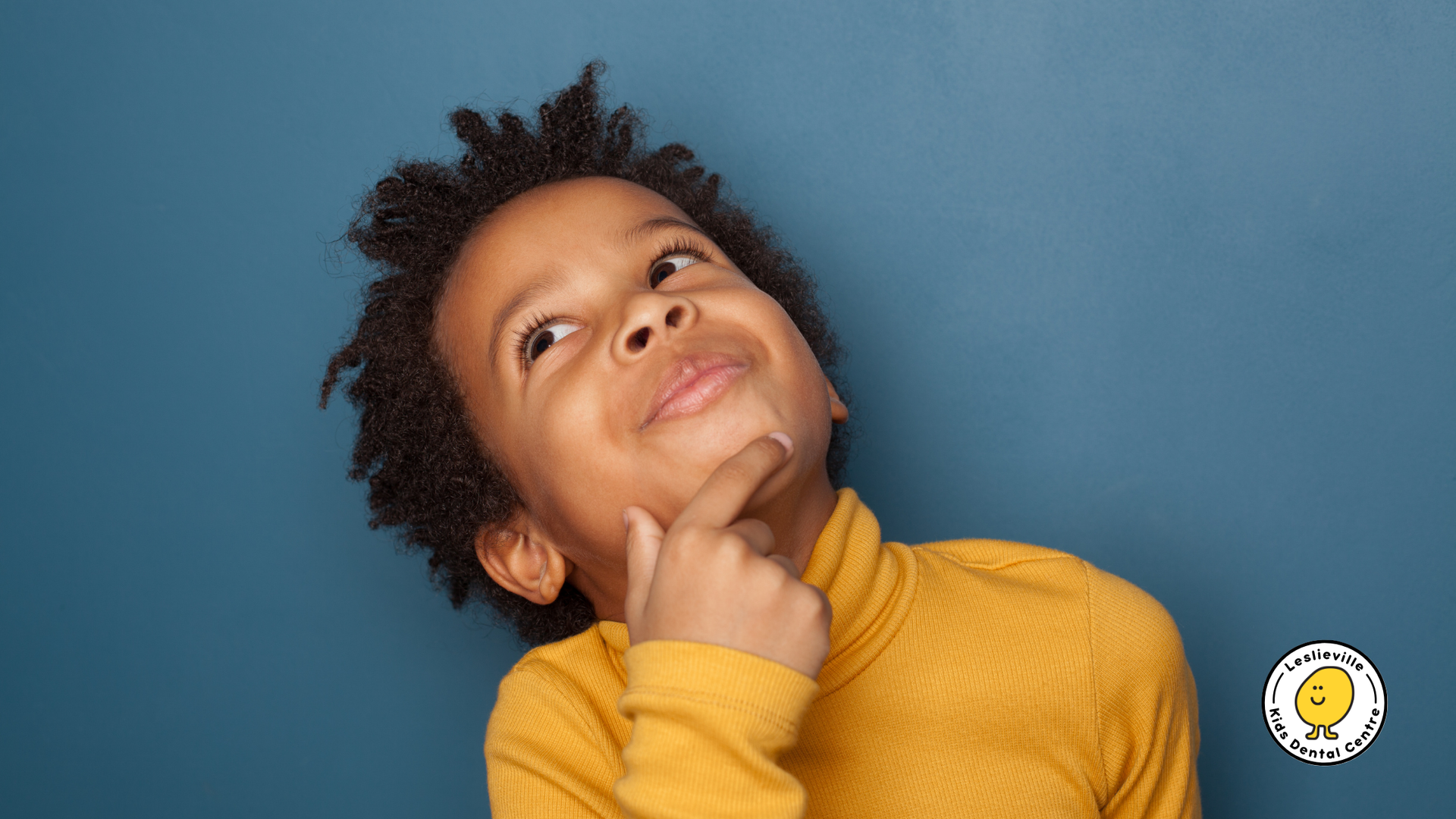 A young boy in a yellow turtleneck is looking up with his hand on his chin.