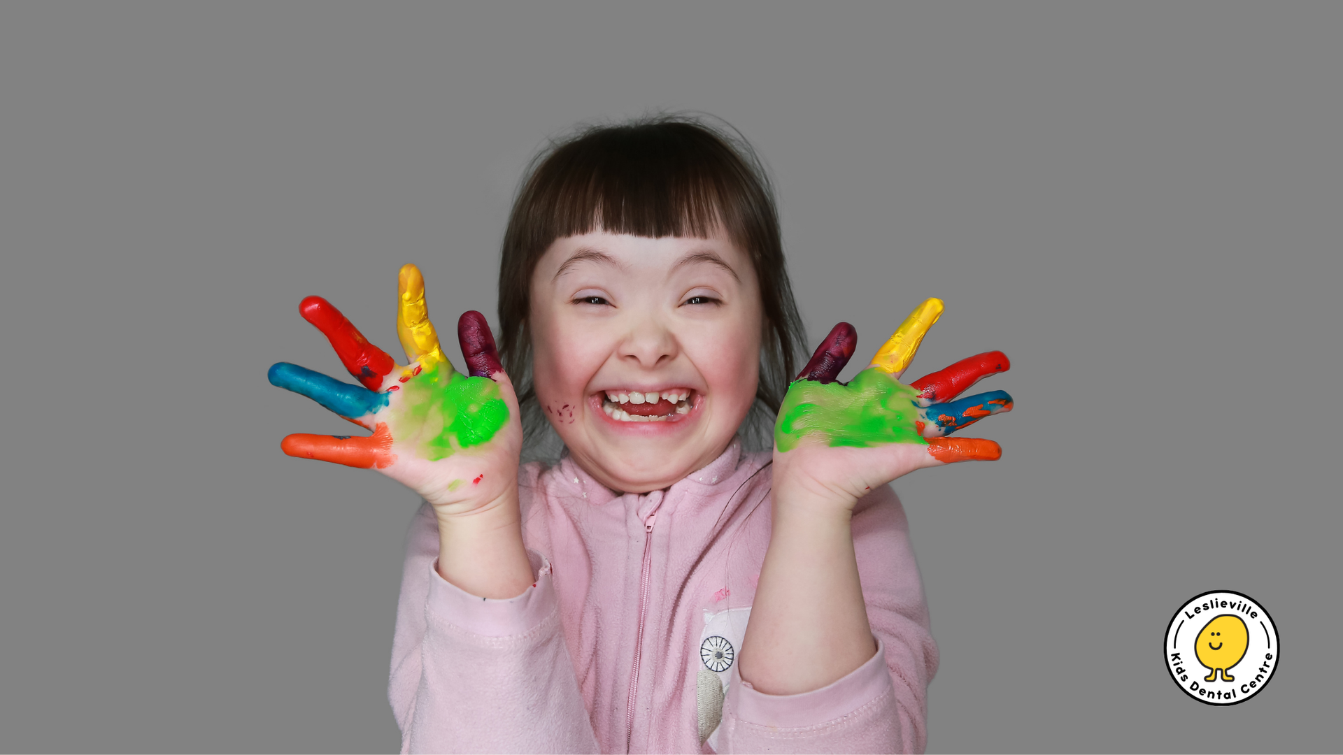Girl with Down syndrome smiling, holding up hands covered in colorful paint. Gray background.
