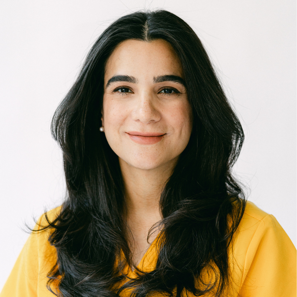 A woman with long black hair is wearing a yellow shirt and smiling.