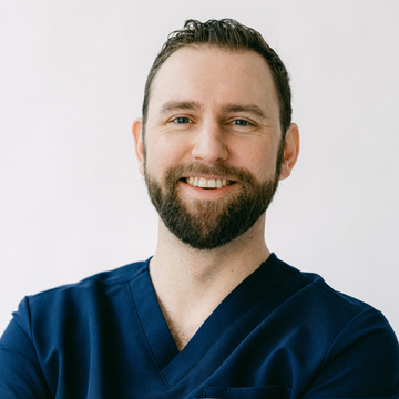 A man with a beard wearing a blue scrub top is smiling for the camera.