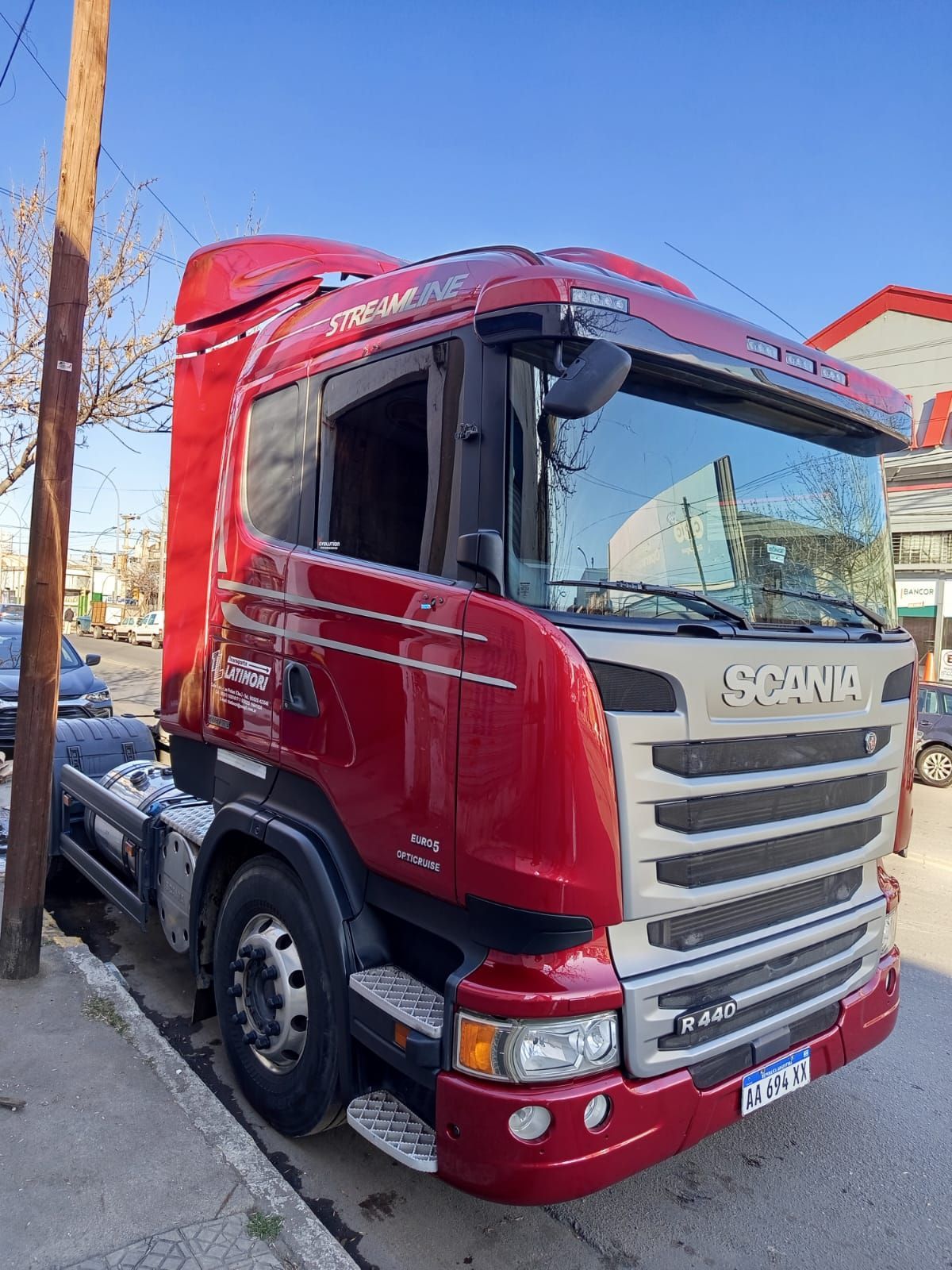 Un camión Scania rojo estacionado en una calle de la ciudad. La luz del sol se refleja en la cabina y se ven algunos coches al fondo.