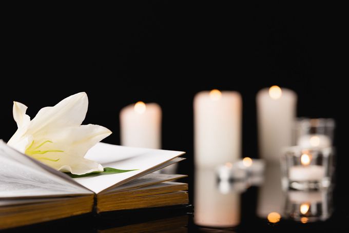 Un fiore bianco poggia su un libro aperto accanto a candele accese in un ambiente buio e riflessivo.