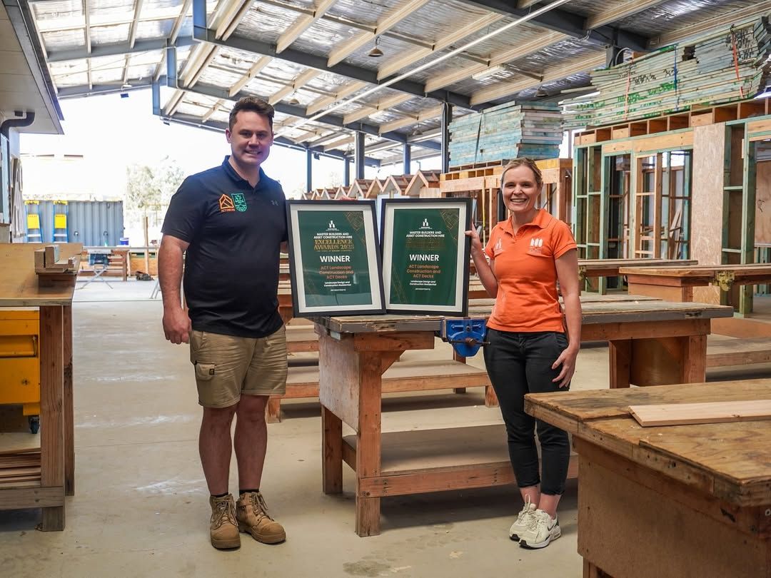Two people in branded shirts stand in a workshop holding two framed award certificates side-by-side on a workbench.