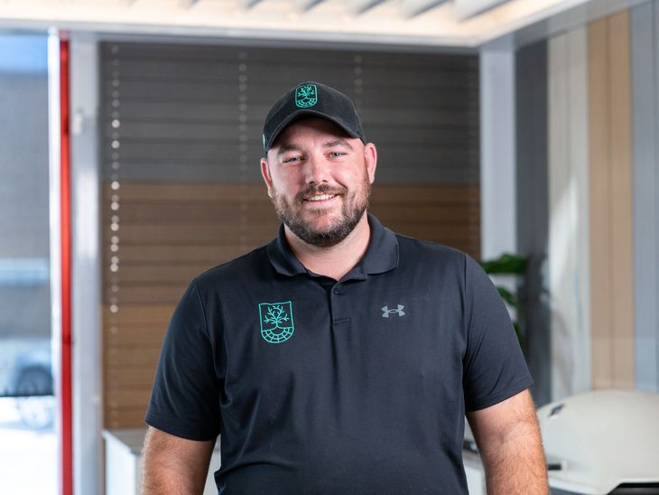 A smiling person with a beard, wearing a black polo and cap with green logos, stands in a modern office with wood accents.