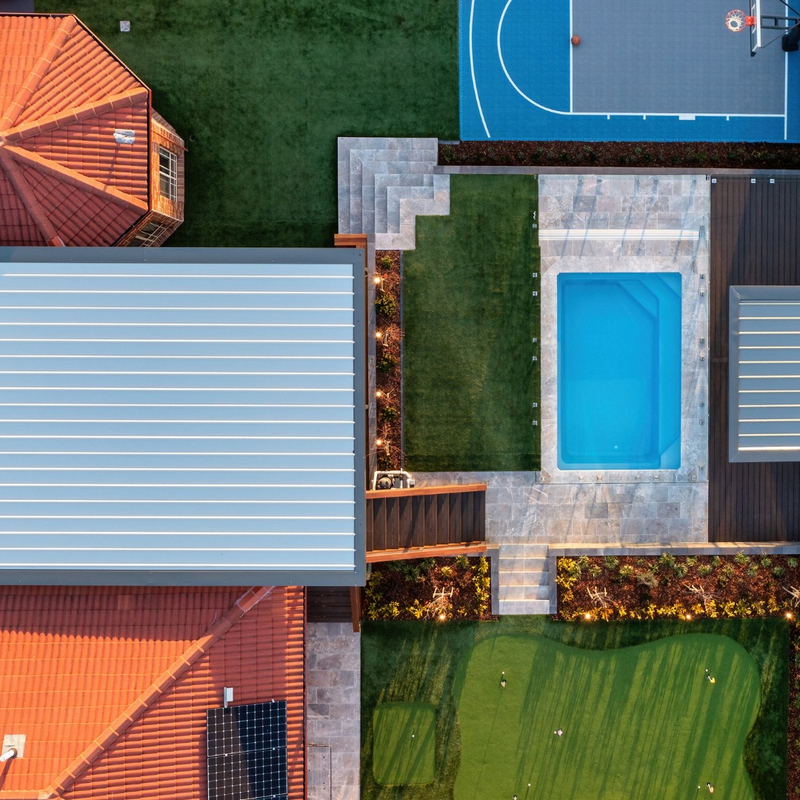 A top-down aerial view of a backyard featuring a swimming pool, basketball court, putting green, and a tiled roof house.