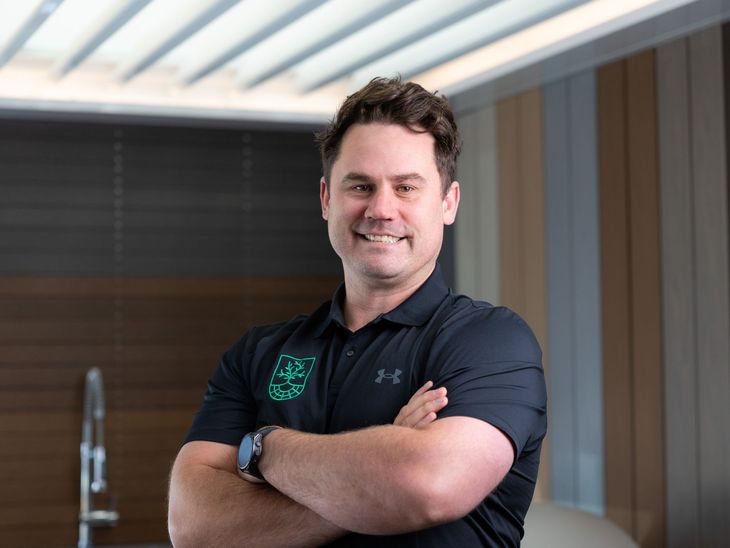 A smiling person with arms crossed, wearing a black Under Armour polo shirt with a green logo, in a modern interior.