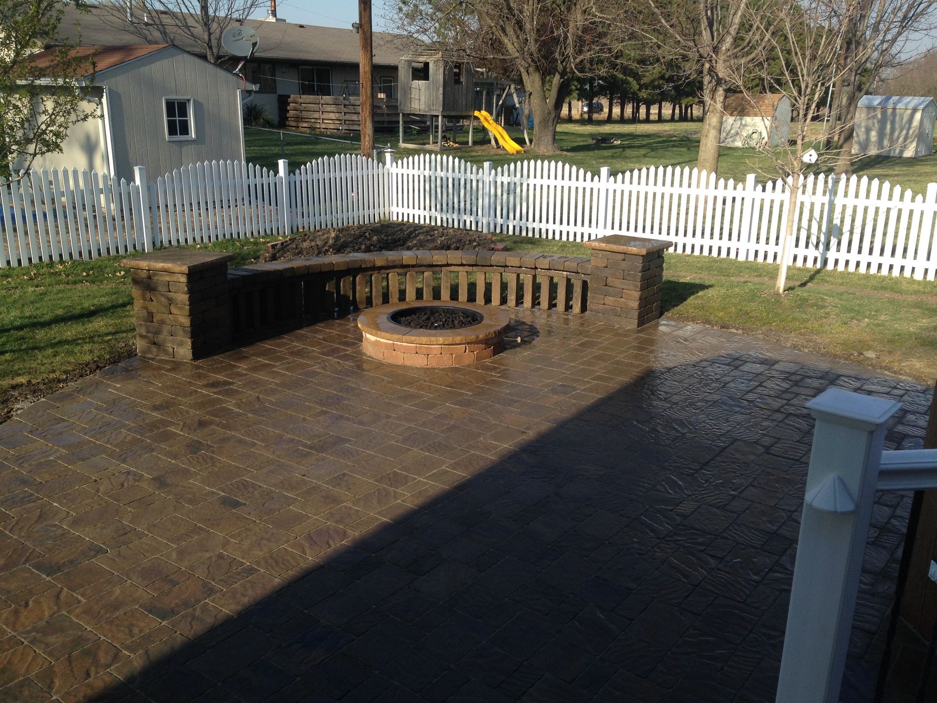 A fire pit is in the middle of a patio next to a white picket fence.