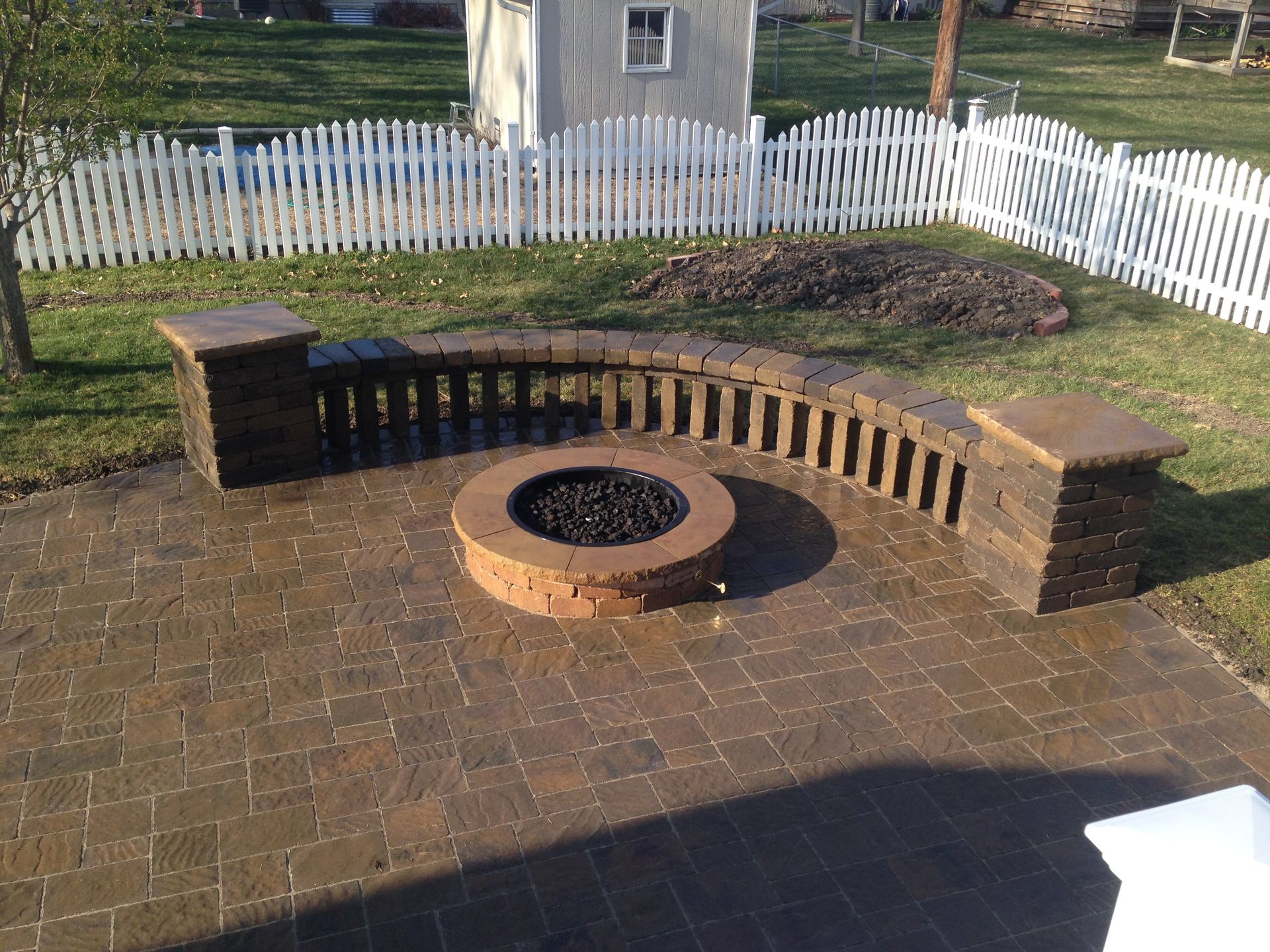 A fire pit on a patio with a white picket fence in the background
