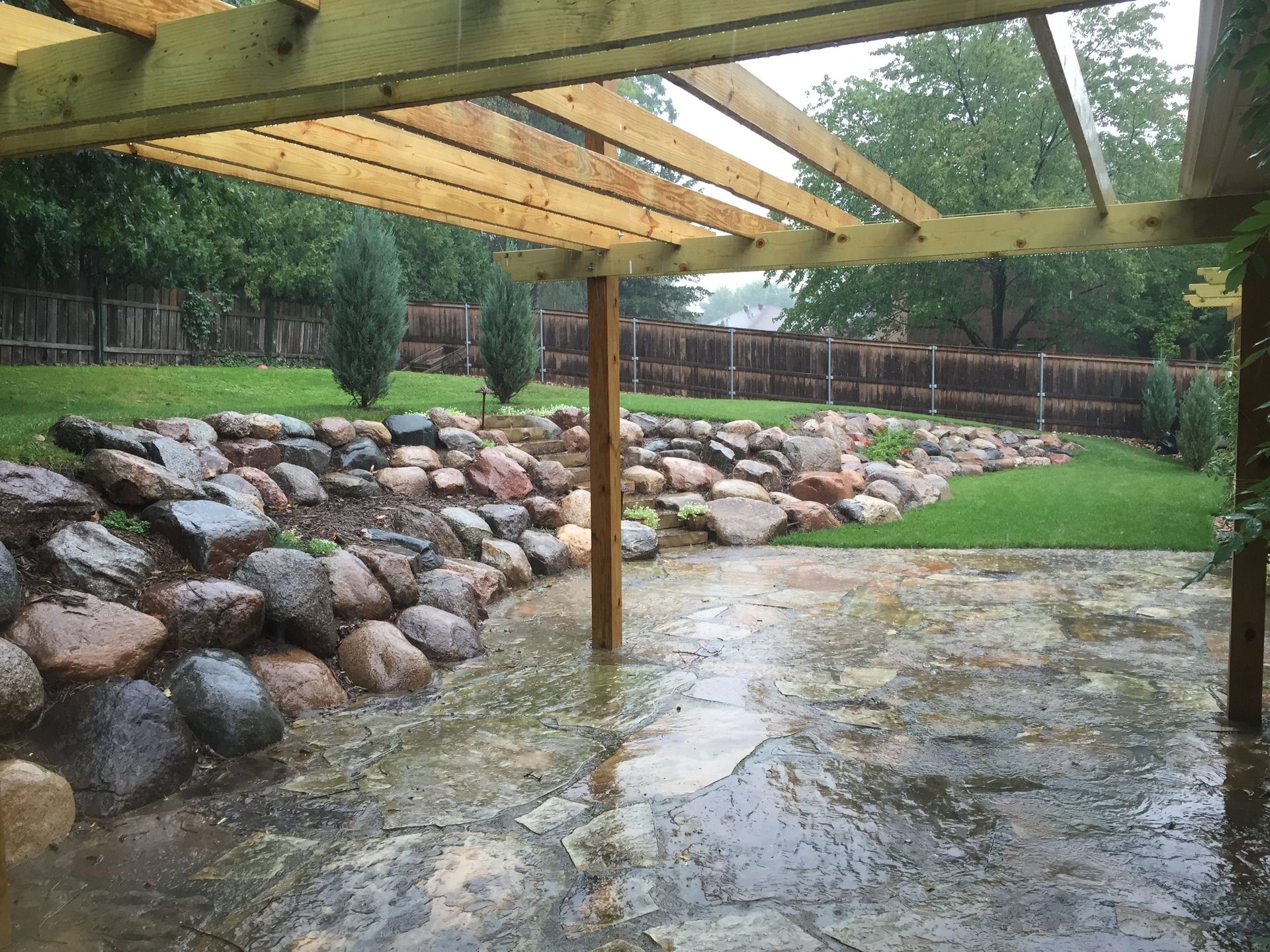 A patio with a wooden pergola and rocks in the background