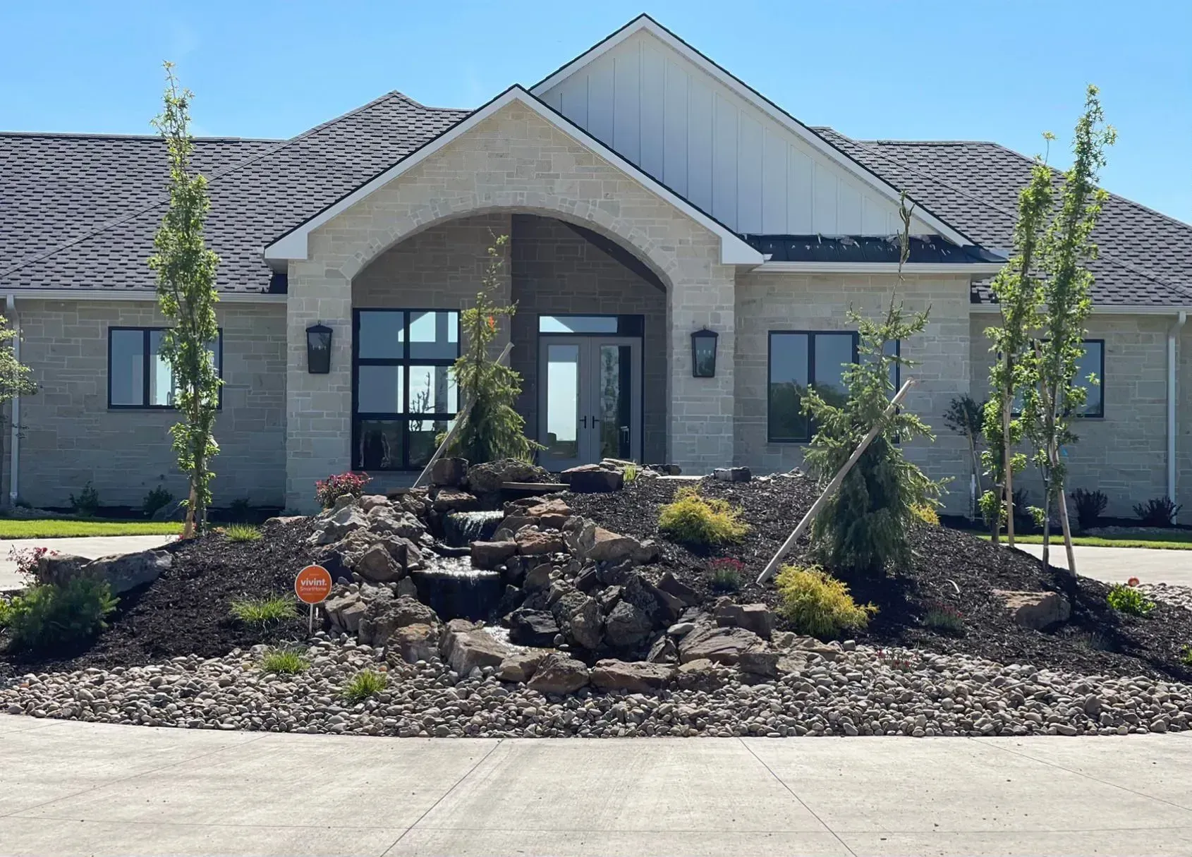 A light-colored stone house with a water feature in front, trees, and gray roof, in a sunny setting.