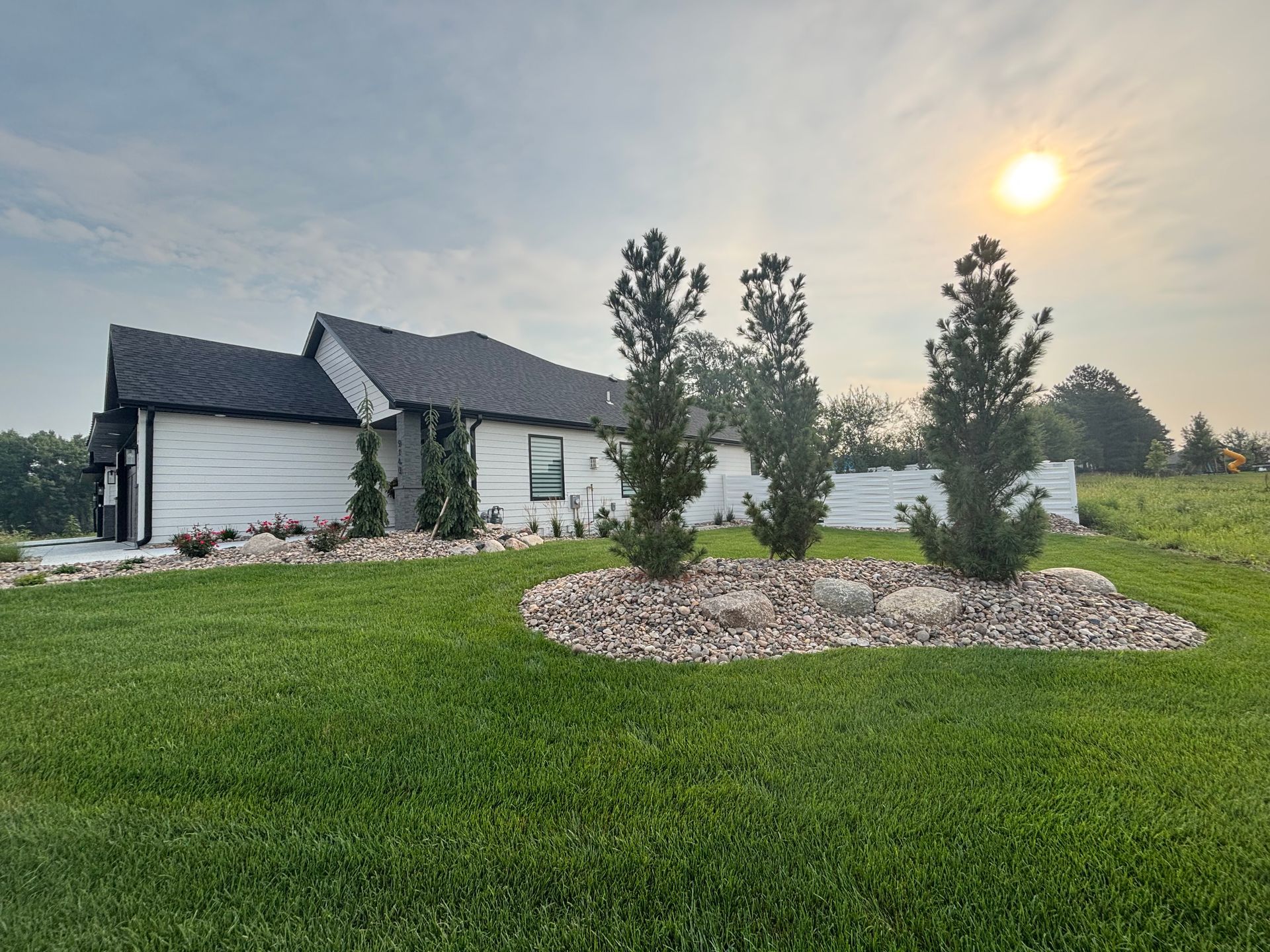 Modern home with green lawn, three trees in a rock garden, and a bright sun in the sky.