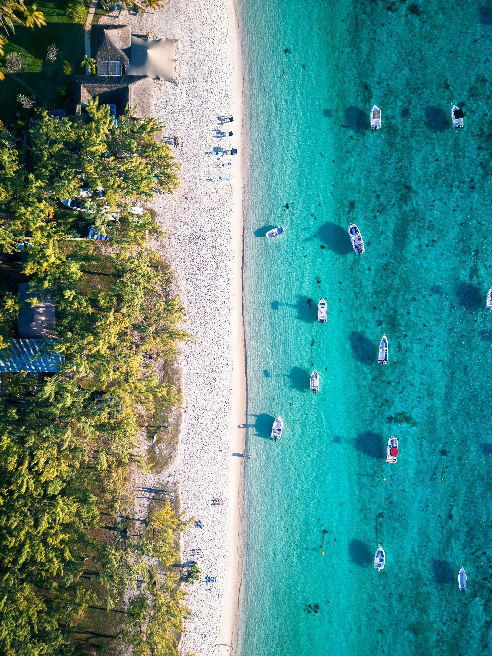 Aerial view of beach with boats in turquoise water and trees on the left.