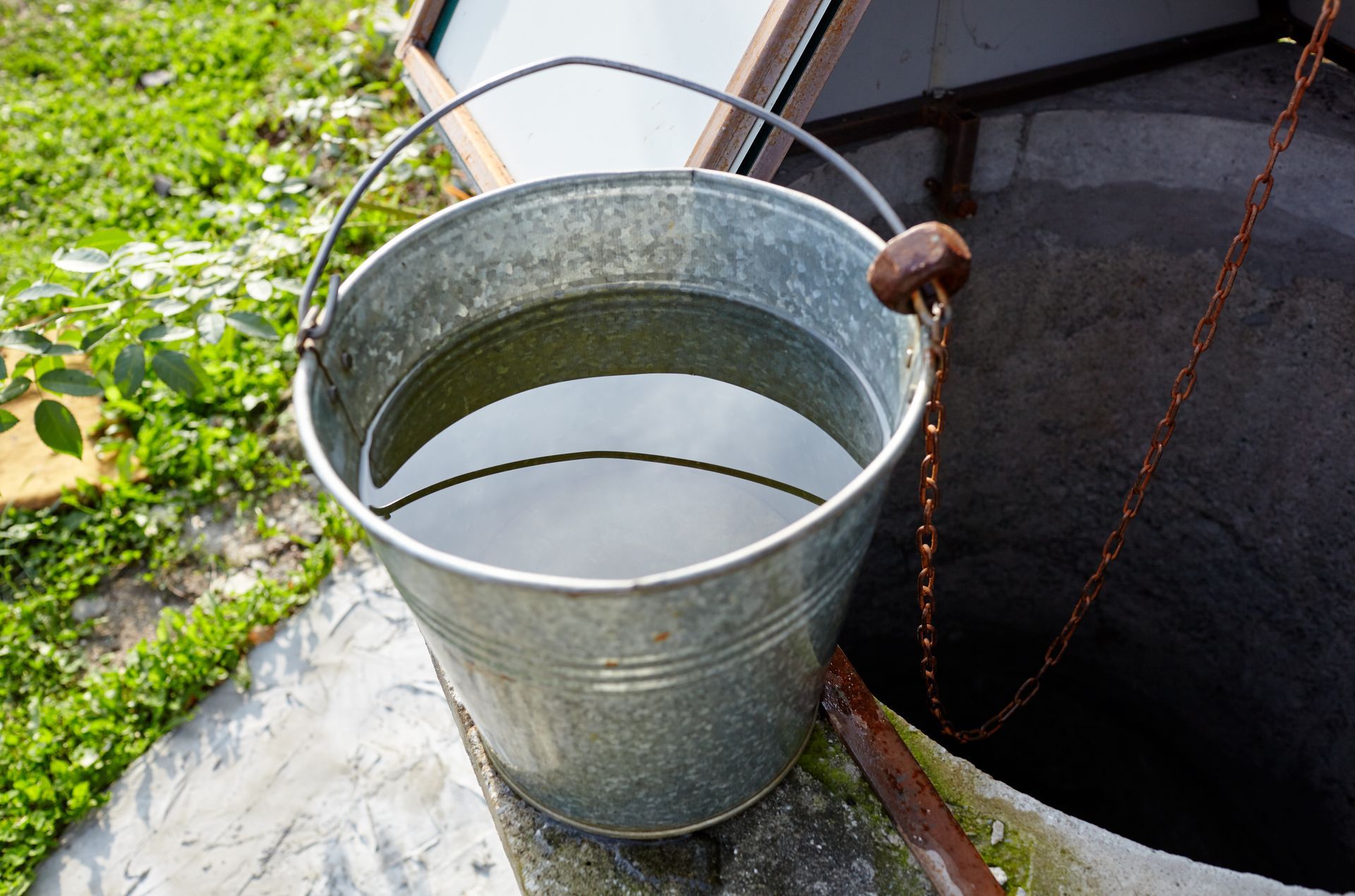 Metal bucket filled with water near an open concrete well with rusty chain.