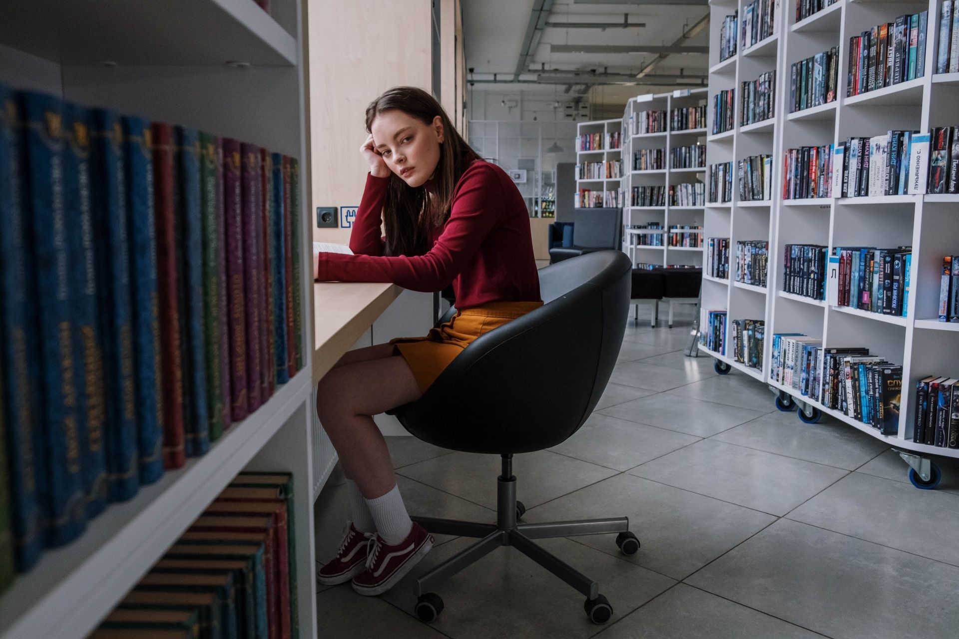 A woman is sitting at a desk in a library surrounded by bookshelves.