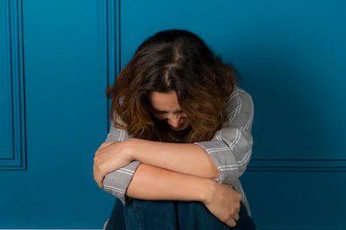 A woman is sitting on the floor hugging her knees against a blue wall.