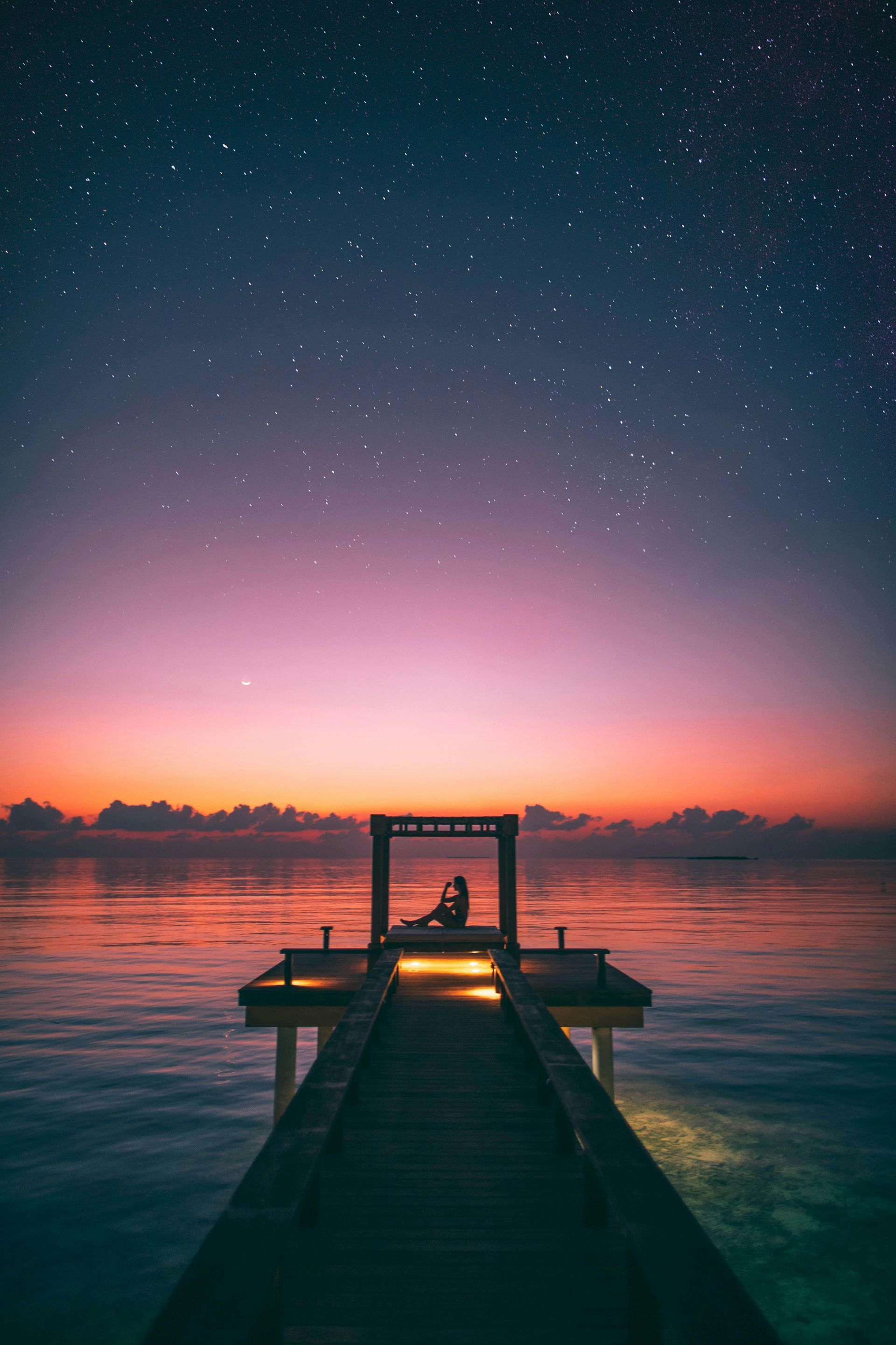 A person is sitting on a dock overlooking the ocean at sunset.