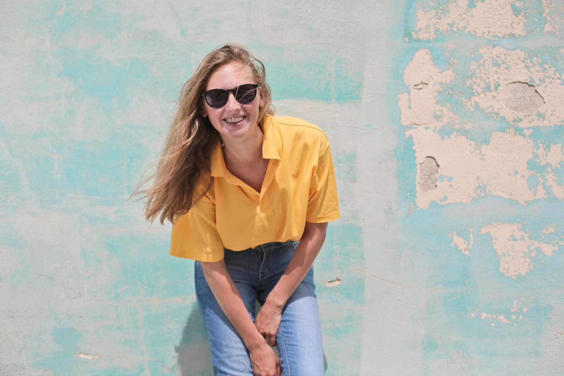 A woman wearing sunglasses and a yellow shirt is sitting in front of a blue wall.