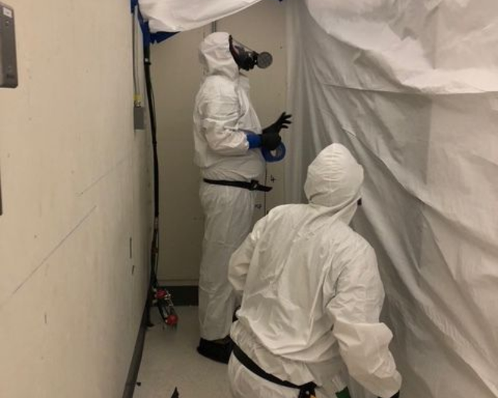 Un homme en combinaison de protection pulvérise une pièce avec un pulvérisateur.