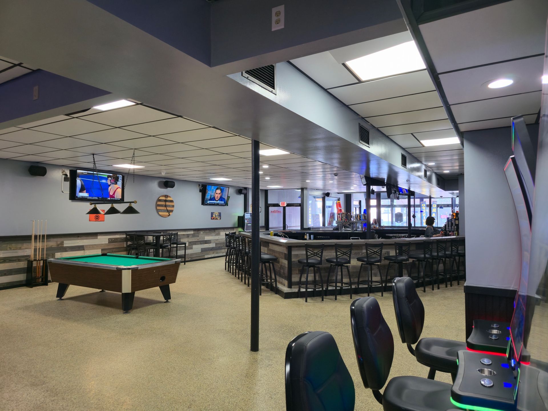 A pool table, bar area with stools, and gaming consoles in a brightly lit, modern recreation room.
