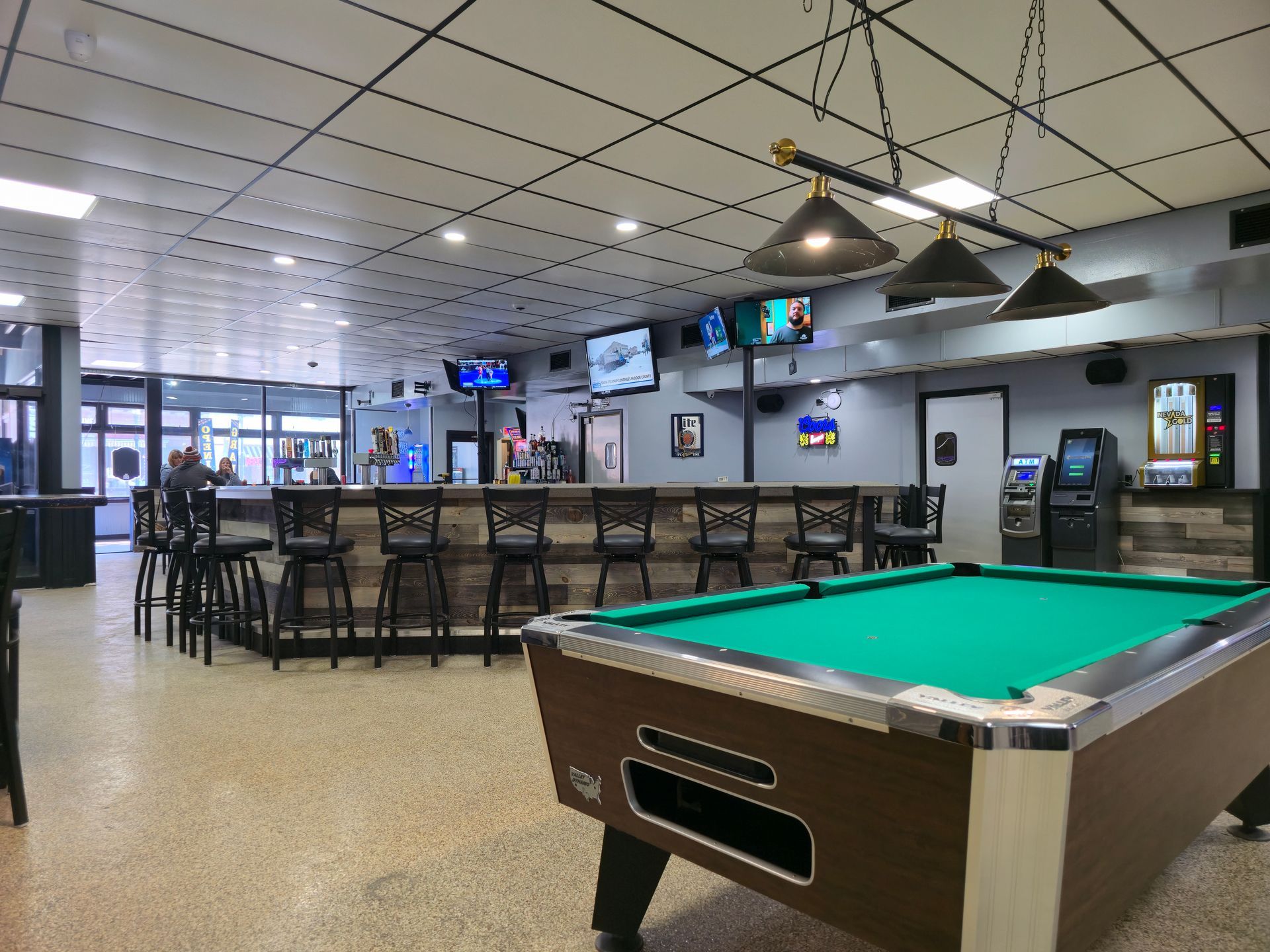 A green felt pool table sits in the foreground of a modern, well-lit tavern with a long bar and stools in the background.